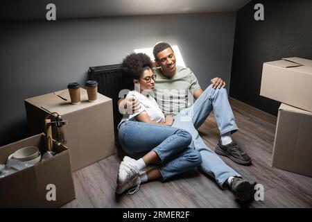 Fröhlich romantisches afroamerikanisches Paar, das sich in der Nähe von Kaffee umarmt, um auf dem Dachboden in einem neuen Haus auf Kisten zu gehen Stockfoto