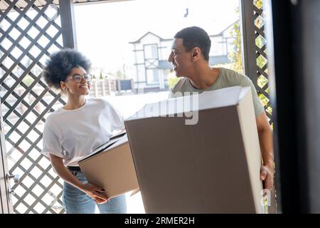 Positives afroamerikanisches Paar, das Kartons in der Nähe der Tür des neuen Hauses während des Umzugs hält Stockfoto
