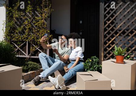 Lächelndes afroamerikanisches Paar mit Kaffee zum Selfie in der Nähe von Kisten und neuem Haus Stockfoto