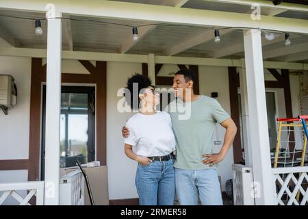 Glückliches afroamerikanisches Paar, das sich umgibt, während es auf einer Veranda in der Nähe eines neuen Hauses im Freien steht Stockfoto