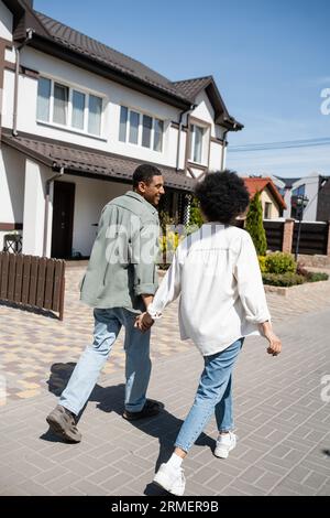 Lächelndes afroamerikanisches Paar, das Hände hält, während es auf dem Bürgersteig in der Nähe von Häusern auf der Straße spaziert Stockfoto