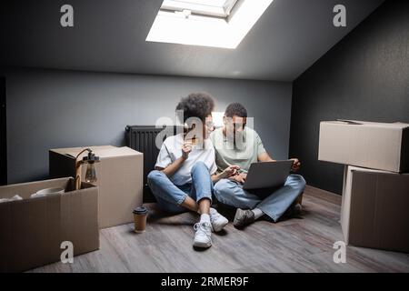 Lächelndes afroamerikanisches Paar mit Laptop und Kreditkarte in der Nähe von Kästen auf dem Dachboden in neuem Haus Stockfoto