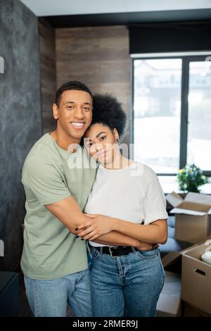 Porträt eines lächelnden afroamerikanischen Paares, das sich in der Nähe verschwommener Kartonschachteln in einem neuen Haus umarmt Stockfoto