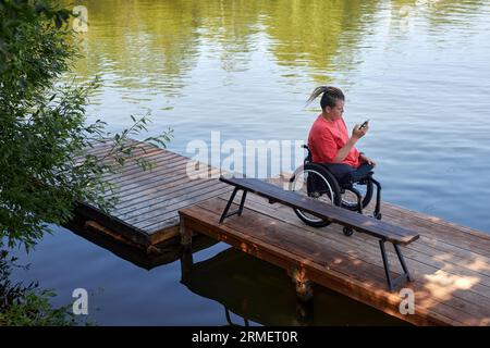 Hochwinkelporträt eines behinderten jungen Mannes, der im Rollstuhl am See sitzt und Smartphone, Kopierraum verwendet Stockfoto