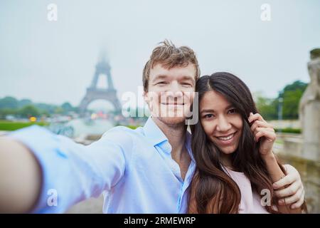 Wunderschönes, romantisches Paar, das sich in der Nähe des Eiffelturms in Paris ein Selfie an einem bewölkten und nebeligen Regentag gönnt Stockfoto