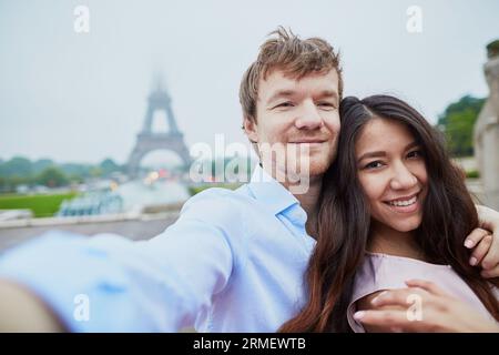 Wunderschönes, romantisches Paar, das sich in der Nähe des Eiffelturms in Paris ein Selfie an einem bewölkten und nebeligen Regentag gönnt Stockfoto