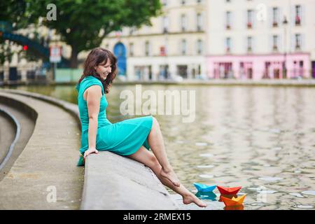 Schöne junge Frau in grünem Kleid spielt mit bunten Papierbooten auf dem Canal Saint Martin in Paris, Frankreich Stockfoto