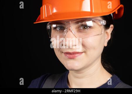 Eine Bauarbeiterin oder Ingenieurin, die eine orangefarbene Schutzmaske und eine Brille auf schwarzem Hintergrund trägt Stockfoto