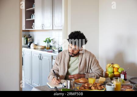 Junger Mann, der frühstückt und einen Planer schreibt Stockfoto