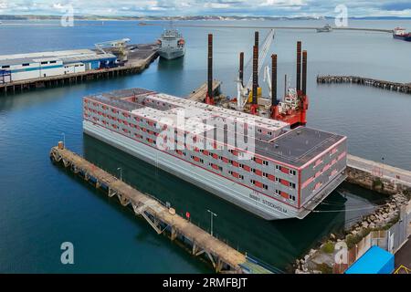 Portland, Dorset, Großbritannien. 28. August 2023. Allgemeine Sicht aus der Luft des leeren Bibby Stockholm Asylbewerberschiffs am Portland Port bei Weymouth in Dorset, der wegen der tödlichen Legionellenbakterien im Wasser evakuiert wurde. Es wird berichtet, dass die Migranten nicht vor Anfang September wieder in den Binnenschiffer einsteigen werden, da sie daran arbeiten, die Legionellenbakterien zu entfernen und zu testen, um festzustellen, ob sie ausgerottet sind. Die Feuerwehrgewerkschaft droht auch rechtliche Schritte, weil sie befürchtet, dass das Schiff unsicher ist. Bildnachweis: Graham Hunt/Alamy Live News Stockfoto
