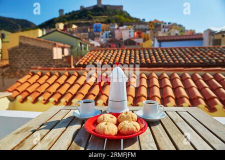 Kaffeemaschine, zwei Tassen frischer Espresso und traditionelle italienische Süßigkeiten auf einem Tisch mit Restaurant, Café oder Terrasse mit Blick auf das Dorf Bosa, Sard Stockfoto