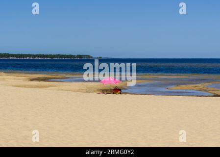 Ein pinkfarbener Sonnenschirm und eine farbige Strandtasche, die am Strand von Arcachon liegen. Klarer Himmel, dunkelblaues Wasser und Strand. Ideal für Hintergrund oder Buchcover. C Stockfoto