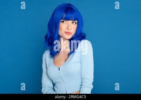 Selbstbewusstes junges weibliches Modell in blauer, heller Perücke und lässiger Kleidung, die träumerisch vor blauem Hintergrund im Studio absieht Stockfoto