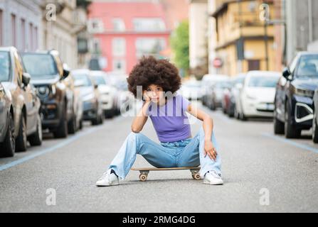 Nachdenkliche junge marokkanische Frau mit Afro-Haaren, die auf die Kamera schaut, während sie auf dem Skateboard auf der Straße in der Nähe von geparkten Autos sitzt, mit Hand berührendem Gesicht und l Stockfoto