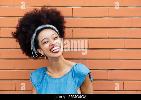 Positive junge afroamerikanische Frau mit Afro-Haar-Stirnband roten Lippen lächelnd und wegschauend, während sie allein gegen braune Ziegelwände steht Stockfoto