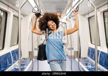 Fröhliche junge gemischte weibliche Passagierin mit dunklem Afro-Haar und roten Lippen, die weit lächeln und in die Kamera schauen, während sie im leeren Zugwagen W steht Stockfoto