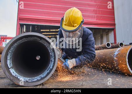 Ein Mann in voller Sicherheitsausrüstung wie Helm, Visier und Weste. Schweißarbeiten an einem rostigen Metallrohr. Ein Mann, der eine tragbare elektrische Säge an einem Metallrohr verwendet. Stockfoto
