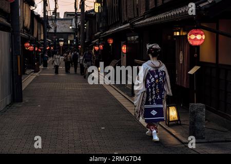 Eine asiatische Frau, die traditionelle japanische Kleidung trägt, einen Kimono, die eine beleuchtete Straße in Kyoto, Japan, entlanggeht Stockfoto