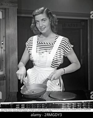 In der Küche der 1940er Eine junge Frau lächelt die Kamera an, die eine Pfanne und einen Schneebesen in den Händen hält. Sie trägt eine weiße Schürze. Schweden 1948. Kristoffersson ref AL27-8 Stockfoto