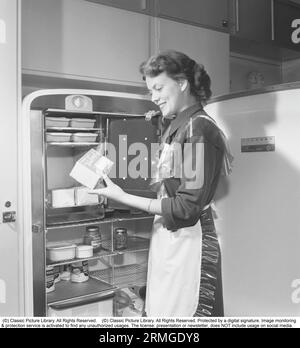 In der Küche der 1950er Das Innere einer Küche und eine junge Frau, die am Leonard-Kühlschrank mit offener Tür steht, zeigt die Dosen und Flaschen mit verschiedenen Lebensmitteln, die auf den Regalen stehen. Ein separater Gefrierschrank ist im Kühlschrank integriert, eine ziemlich neue Funktion zu diesem Zeitpunkt. Sie trägt eine Plastikschürze. Sie ist Haide Göransson, 1928–2008, Modemodel und Schauspielerin. Schweden 1950 Kristoffersson ref AU22-10 Stockfoto