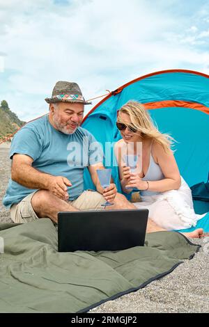 Das Paar hat Spaß, trinkt Wein und jubelt dem Spielvideo auf dem Laptop beim Picknick in der Nähe des Zeltes am Meer zu. Sommer Strandurlaub im Freien. Stockfoto