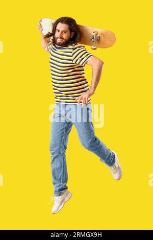 Springender bärtiger junger Mann mit Skateboard auf gelbem Hintergrund Stockfoto