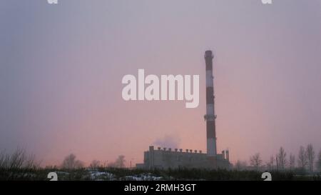 Der Kamin und das Gebäude des Kesselhauses im Nebel während eines Sturms vor dem Hintergrund des Lichts der Gewächshäuser Stockfoto