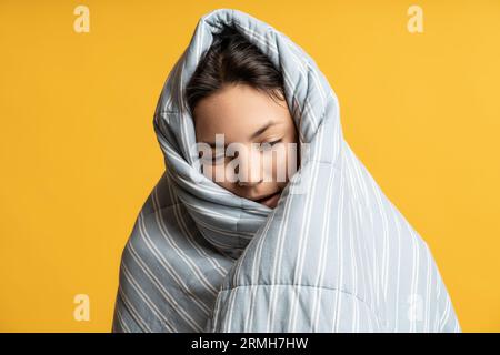 Porträt einer verschlafenen brünetten Frau, die in eine graue Decke auf gelbem Hintergrund gehüllt ist. Stockfoto