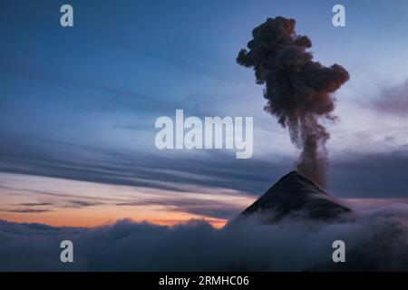 Eine dramatische Szene, wie der Fuego-Vulkan in Guatemala durch eine niedrige Abendwolke ausbricht und eine dunkle Aschewolke hoch in die Luft schickt Stockfoto