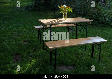 Einfacher Holztisch mit Blumenstrauß und Bänken auf der Wiese in einem natürlichen Garten an einem sonnigen Tag, idyllische Sommerszene, Kopierraum, ausgewählte Auswahl Stockfoto