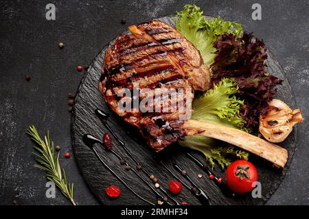 Tomahawk Steak auf den Knochen, mit Kräutern, Tomaten und Knoblauch, auf schwarzem Schiefer, auf dunklem Beton Stockfoto