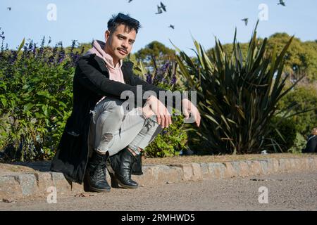 Junger argentinischer lateinamerikaner aus der lgbt-Gemeinschaft, mit schwarzem Pullover, Hose und schwarzen Lederstiefeln, sitzt im öffentlichen Park und schaut auf die Kamera. Stockfoto