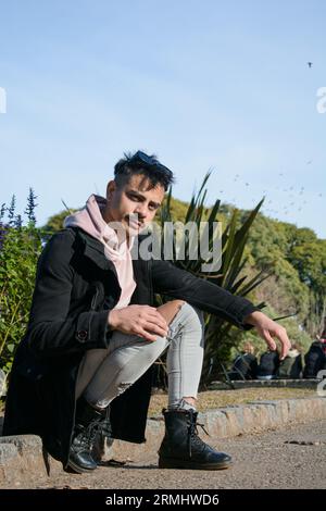 Junger argentinischer lateinamerikaner aus der lgbt-Gemeinschaft, mit schwarzem Pullover, Hose und schwarzen Lederstiefeln, sitzt im öffentlichen Park und schaut auf die Kamera. Stockfoto