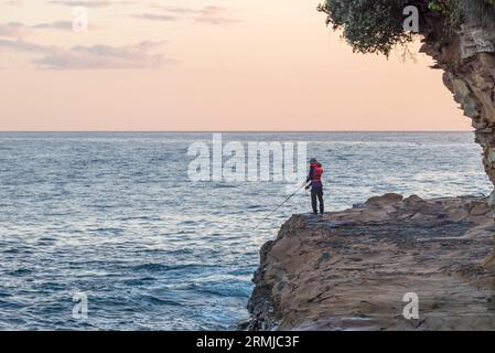 Ein Mann mit einer Schwimmweste steht am beliebten südlichen Ende von Avoca Beach an der Central Coast von New South Wales, Australien, zum Angeln an den Felsen Stockfoto