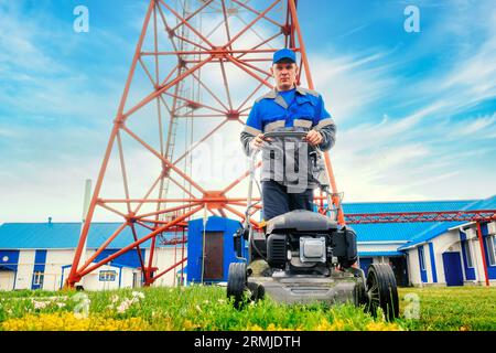 Arbeiter im Overall mäht Gebiet der Industrieanlage mit Rasenmäher. Der Handwerker geht mit dem Rasenmäher gegen den blauen Himmel. Stockfoto