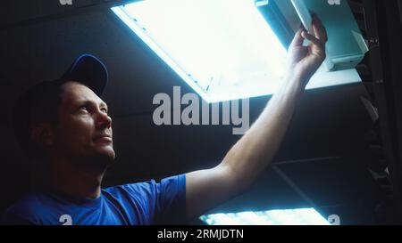 Professioneller Elektriker wechselt Leuchtstofflampe in Deckenlampe. Arbeiter repariert Beleuchtung im Bürogebäude. Ansicht von unten nach oben. Elektriker bei der Arbeit. Echte Arbeitsabläufe. Stockfoto
