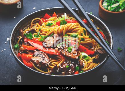 Bratnudeln mit Gemüse umrühren: Rote Paprika, Champignons, grüne Zwiebel und Sesamsamen in Keramikschüssel. Schwarzer Tischhintergrund, Draufsicht Stockfoto