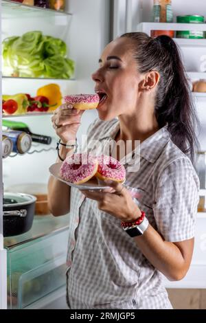 Hungrige Brünette im Pyjama genießt süße Donuts spät in der Nacht am offenen Kühlschrank. Stockfoto