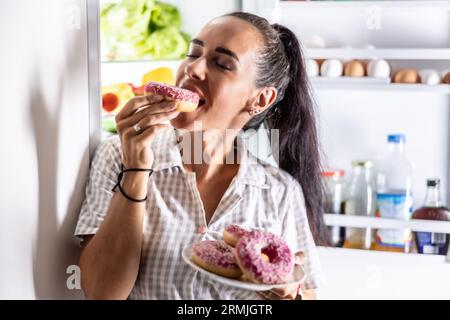 Hungrige Brünette im Pyjama genießt süße Donuts spät in der Nacht am offenen Kühlschrank. Stockfoto