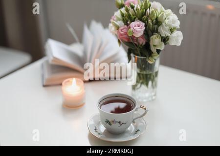 Romantisches Stillleben, ein Rosenstrauß in einer Vase, eine Tasse Tee und ein Buch auf weißem Tisch Stockfoto