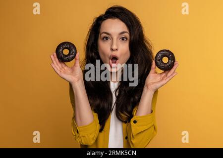 Niedliche Brünette posiert für die Kamera mit köstlichen Keksen auf dem gelben Hintergrund während des Fotoshootings im Studio. Ungesunde Ernährung Konzept Stockfoto