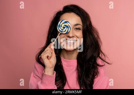 Lächelnde glückliche junge Frau posiert mit einem köstlichen Bonbon in der Hand auf dem rosa Hintergrund. Süßzahnkonzept Stockfoto