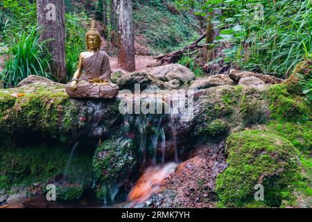 Buddha-Statue sitzt in einem Gebirgsfluss und meditiert auf einem Wasserfall mit grünem Moos und Felsen Stockfoto