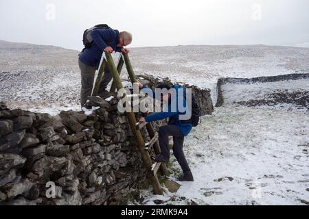 Zwei ältere Männer (Wanderer) klettern Holzleiter Stile in Snow over Dry Stone Wall in der Nähe von Austwick im Yorkshire Dales National Park, England, Großbritannien. Stockfoto