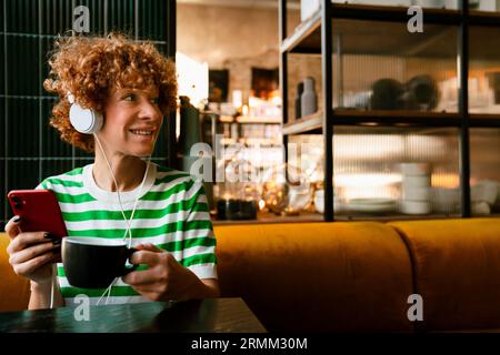 Mittelweiße Frau mit lockigem Haar, die Handy und Kopfhörer benutzt, während sie am Tisch im Café sitzt Stockfoto