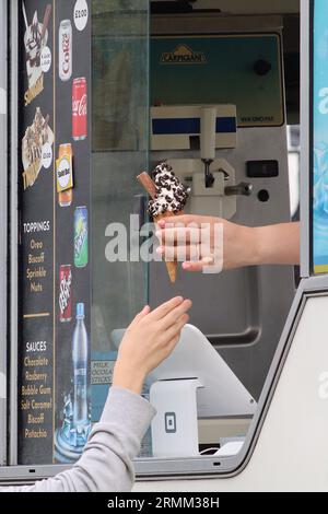 Ein Eisverkäufer gibt im August 2023 ein Eis aus dem Jahr 99 mit Schokoladenchips aus der Servierluke an einen wartenden Kunden weiter. Stockfoto