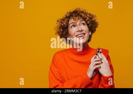 Nachdenkliche Ingwerfrau mittleren Alters, die Smartphone hält, während sie isoliert über der gelben Wand steht Stockfoto