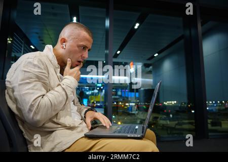 Der junge Erwachsene kaukasier sieht überrascht aus, als er in der Flughafenlounge an einem Laptop arbeitet. Schockierter und besorgter Mann liest Nachrichten auf dem Laptop. Verwirrter Freiberufler Stockfoto