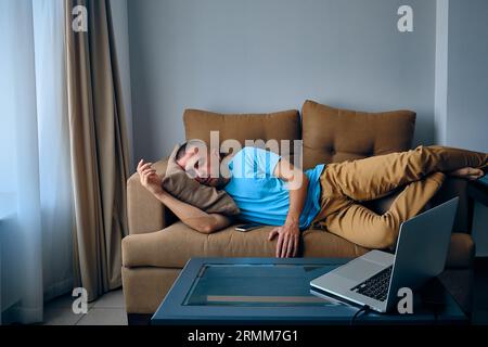 Junger, erwachsener kaukasier schlief auf einer Couch und schaute sich Filme auf dem Laptop an. Müder Mann schläft auf dem Sofa Stockfoto