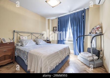 Ein Doppelzimmer im klassischen Stil mit einem Kopfteil aus Metall auf dem Bett und blauen Vorhängen auf dem Fenster. Stockfoto